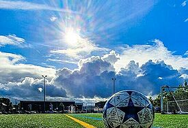 Ein schmutziger Fußball liegt auf einem Fußballplatz im grünen Gras, am Himmel zwischen leichten weißen Wolkenschleiern scheint die Sonne. Im Hintergrund stehen Menschen auf dem Spielfeld.