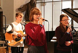 Drei junge Menschen auf der Bühne, ein junger Mann spielt Gitarre, zwei Frauen singen.