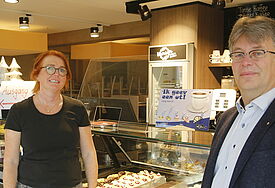 Pastor Raabe und Doris Hansen stehen vor einem gut gefüllten Tresen in einer Bäckerei. Darauf steht ein Schild mit der Aufschrift "Ik geev een ut – lang man bi". 