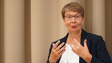 Landesbischöfin Kristina Kühnbaum-Schmidt gestikuliert mit ihrern Händen. Sie sitzt vor einem Vorhang in beige.