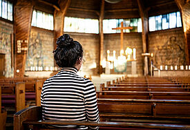 Das Bild zeigt Fofo, eine junge Frau aus dem Irak, in der Matthäuskirche in Darmstadt. Sie sitzt in der Kirche, ihre Haare sind zu einem Knoten hochgesteckt. Ihr Gesicht ist nicht zu erkennen.