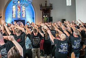 Innenraum der Kirche, in der Menschen in schwarzer Kleidung kollektiv die Arme in die Höhe recken