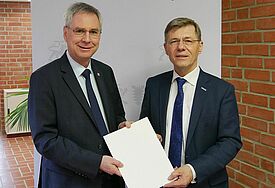 Zwei Männer im Anzug in einem Büroflur. Sie halten ein Papier in der Hand