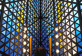 Kirchraum von innen, die Wand besteht aus bunten Glasdreiecken, durch die das Licht fällt; in der Mitte ist der Altar mit zwei Kerzen