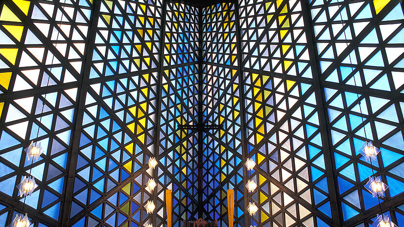 Kirchraum von innen, die Wand besteht aus bunten Glasdreiecken, durch die das Licht fällt; in der Mitte ist der Altar mit zwei Kerzen