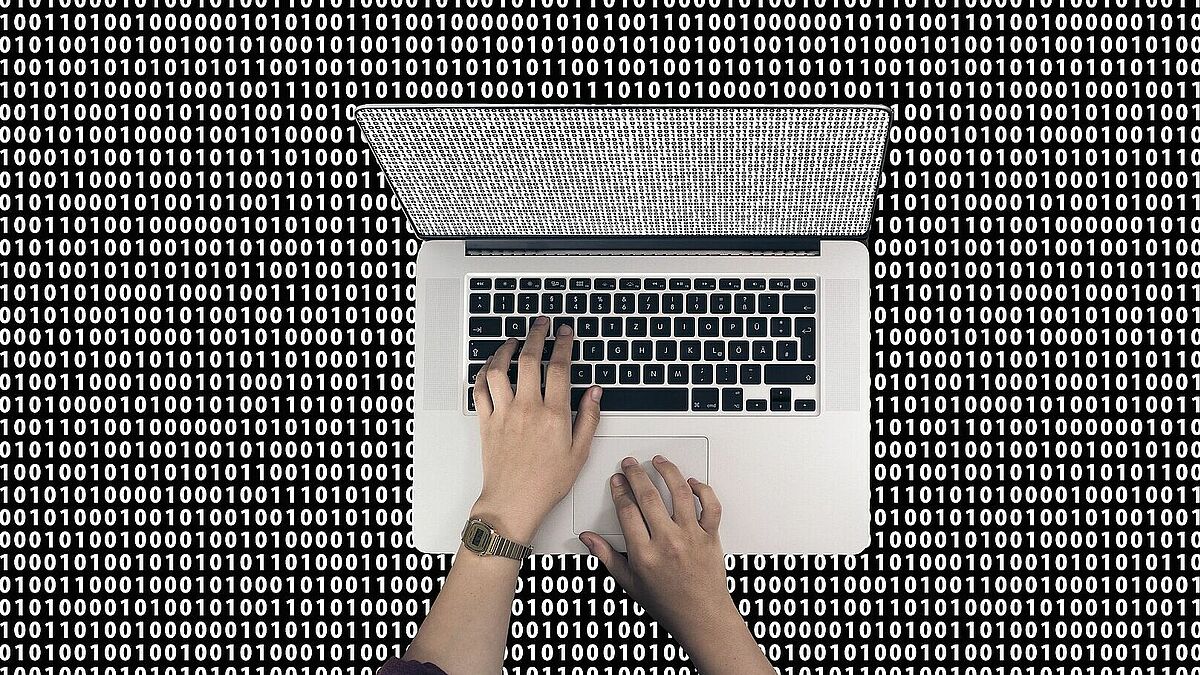 Laptop von oben aufgenommen, auf der Tastaur zwei tippende Hände, drumherum Nullen und Einsen
