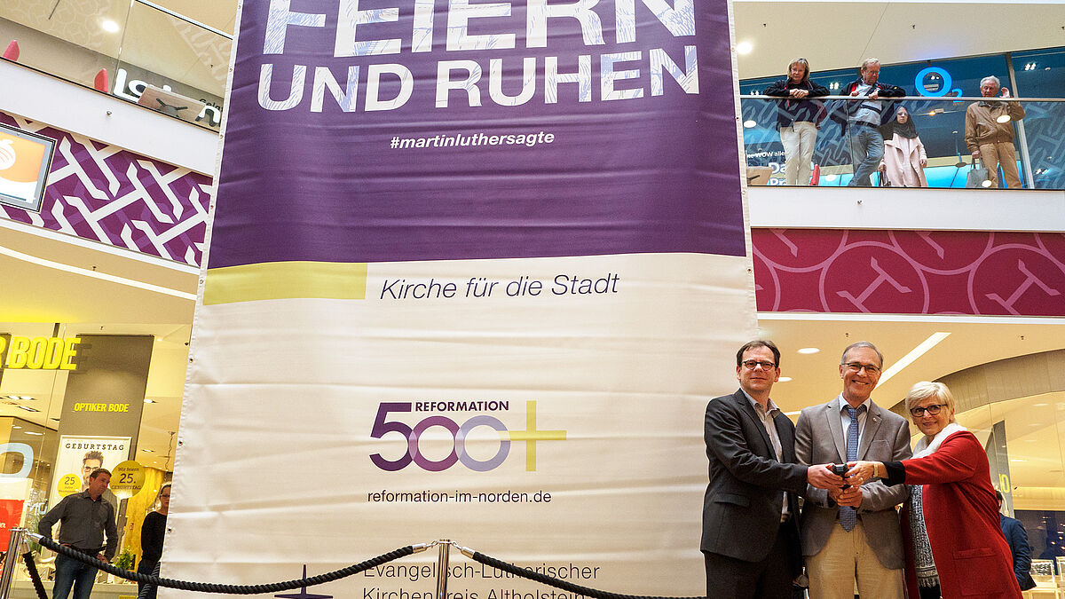 Oberbürgermeister Dr. Olaf Tauras, Propst Stefan Block und Stadtpräsidentin Anna-Katharina Schättiger lassen das Martin-Luther-Zitat "Man kann Gott nicht allein mit Arbeit dienen, sondern auch mit Feiern und Ruhen" an seinen Platz in der Holstengalerie hochziehen. 