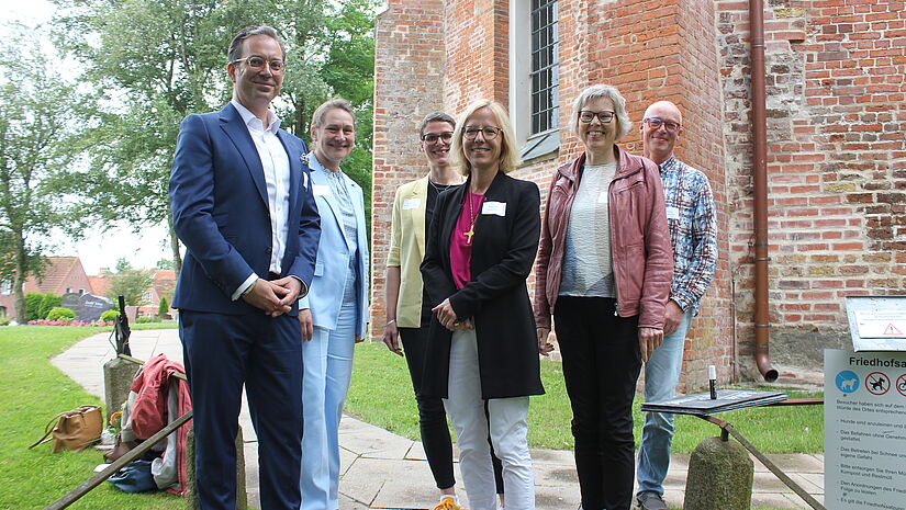 Sechs Menschen stehen vor einer alten Kirchenmauer aus Backstein und blicken in die Kamera