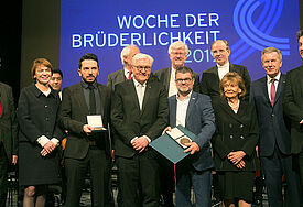 Gruppe an Menschen in Anzügen und Kostümen vor einer Projektion mit dem Schriftzug "Woche der Brüderlichkeit"