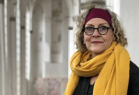 Frau trägt einen gelben Schal und ein rotes Stirnband, schaut in die Kamera, im Hintergrund die gotischen Bögen der St. Petri-Kirche