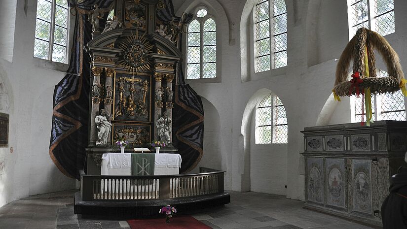 Weiß gestrichener Kirchraum mit Mosaikfenstern, in der Mitte der Altar