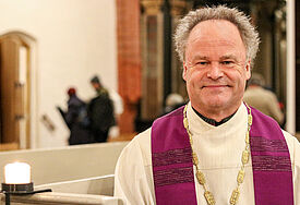 Bischof Tilman Jeremias sitzt in der Kirchenbank der Greifswalder St. Marienkirche, eine Kerze brennt, er trägt eine violette Stola 