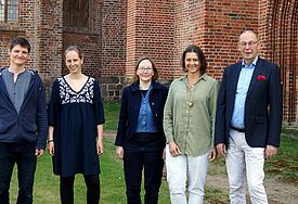 Zwei Männer und drei Frauen stehen auf grünem Rasen vor einer Kirche aus rotem Backstein.