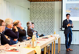 Links sitzen Leute am Tisch, rechts steht der Referent vor einer Leinwand mit einer Präsentation und redet