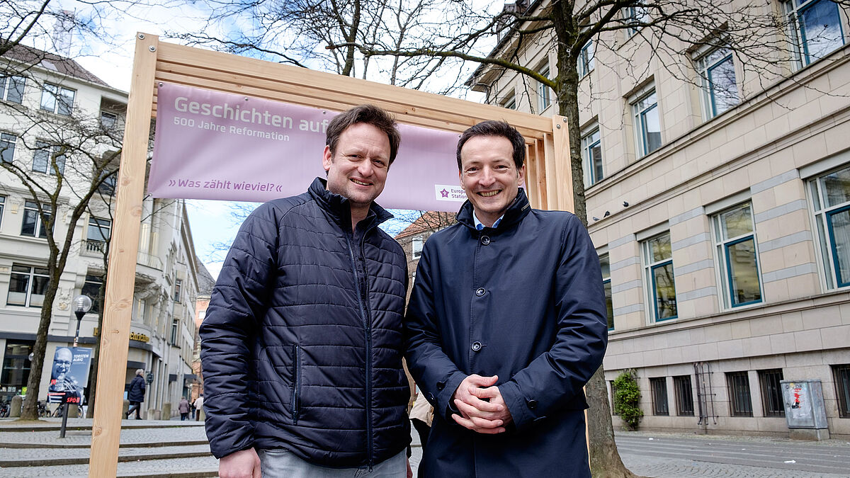 Wilko Teifke (Kirchenkreis Altholstein, v.l.) und Daniel Mourkojannis (Nordkirche) vor dem Tor in der Kieler Fußgängerzone, das zum Europäischen Stationenweg einlädt.