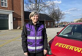Eine Frau mit einer violetten Weste steht neben einem kleinen roten Auto mit der Aufschrift Feuerwehr.