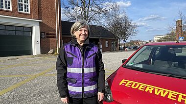 Eine Frau mit einer violetten Weste steht neben einem kleinen roten Auto mit der Aufschrift Feuerwehr.