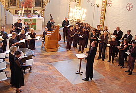Menschen in schwarzer Kleidung mit Text und Noten in der Hand stehen in einem Halbkreis vor dem Altar und singen unter der Leitung eines Chorleiters, der mit Notenpult in der Mitte steht.