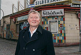 Pastor Sieghart Wilm steht mit schwarzer Jacke vor einer Häuserecke. Im Hintergrund ist der Schriftzug "Zum Silbersack" am Gebäude zu lesen.