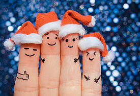 Vier Finger einer Hand tragen Weihnachtsmützen und haben lustige aufgemalte Gesichter, im Hintergrund glitzert es.