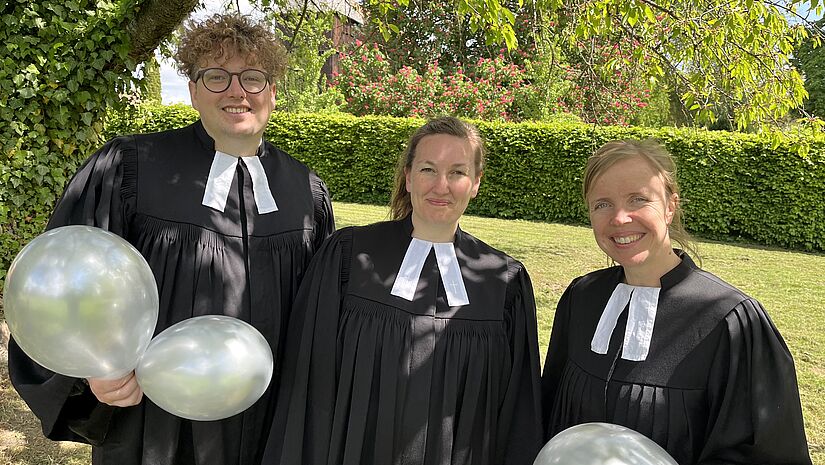 Zwei Frauen und ein Mann stehen im Talar vor einer grünen Rasenfläche und halten weiße Luftballons in den Händen.