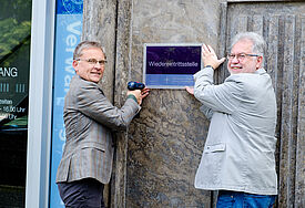 Propst Thomas Lienau-Becker (l.) und Verwaltungsleiter Ralf Stolte bringen das Schild der Wiedereintrittsstelle am Martensdamm 2 in Kiel an.
