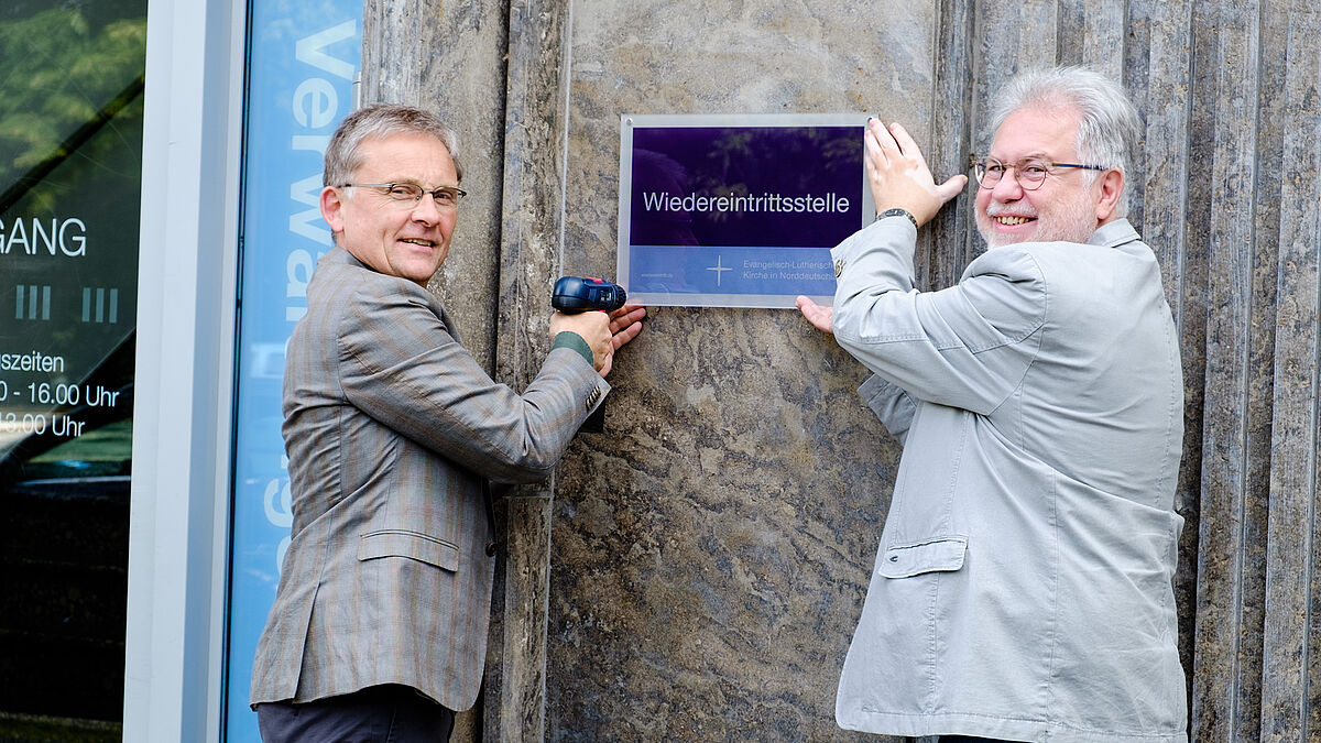 Propst Thomas Lienau-Becker (l.) und Verwaltungsleiter Ralf Stolte bringen das Schild der Wiedereintrittsstelle am Martensdamm 2 in Kiel an.
