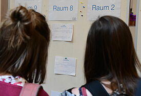 Zwei Köpfe von hinten unscharf im Vordergrund, in der Mitte ist der Sessionplan an der Pinnwand zu sehen