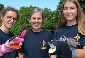 Drei Frauen lachen in die Kamera, links und rechts werden bunte Handpuppen ins Bild gehalten