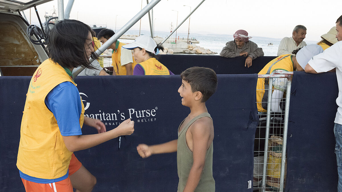 Chios, Greece - July 21, 2016: A female Asian volunteer from the People's Street Kitchen (PSK) and a refugee boy are playing rock-paper-scissors in the Souda Refugee Camp on the Island of Chios in Greece on a hot summer evening. The volunteer has just finished serving dinner for the refugees together with other PSK volunteers, Norwegian NGO Drop in the Ocean volunteers and Samaritan's Purse officials.