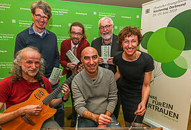 Sechs Menschen vor der grünen Kirchentagswand, links das Kirchentagsmotiv mit Luftballons und schwebendem Menschen), alle lachen in die Kamera, zwei Menschen vorn mit Saiteninstrumenten)