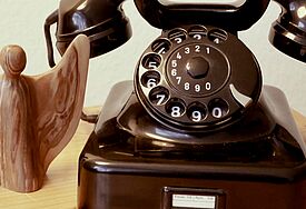 Ein altes, schwarzes Telefon mit Wählscheibe steht neben einem Engel aus Holz.