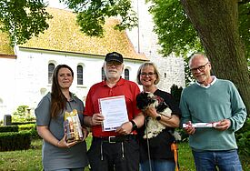 Küster Michael Hirner nimmt die Auszeichnung "ökofaire Gemeinde der Nordkirche" entgegen. Auf dem Foto zu sehen sind vor der Kirche St. Petri Bosau ebenso: Elena Stern vom Kirchenkreis (links), die stellvertretende Kirchengemeinderatsvorsitzende Sabine Hirner mit Hund Fine und Tobias Jahn vom Küsterarbeitskreis (rechts im Bild). 