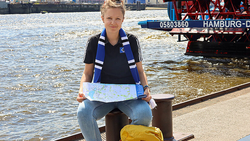 Anna Stöcken sitzt auf einem Poller an der Elbe. In den Händen hält sie eine aufgeklappte Karte. Umihren Hals hängt ein blau-schwarz-weißer HAS-Fanschal.