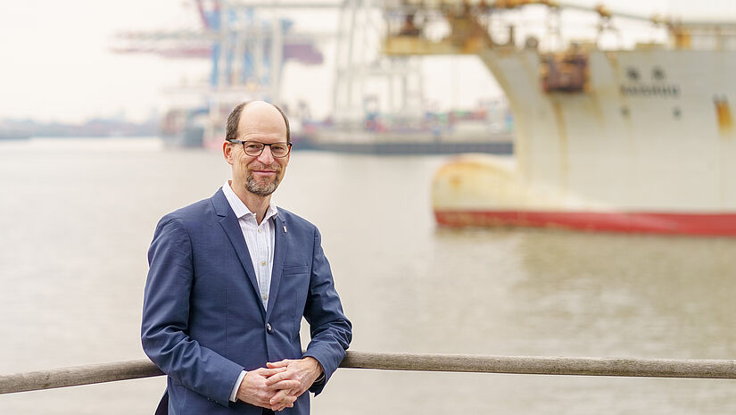 Matthias Ristau steht an ein Geländer gelehnt am Ufer der Elbe in Hamburg Altona. Im Hintergrund ist ein Schiff zu sehen.