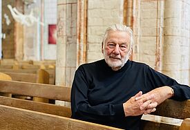 Stefan Schmidt sitzt in einer Kirchenbank. Er trägt einen dunklen, hoch geschlossenen Pullover und blickt direkt in die Kamera.