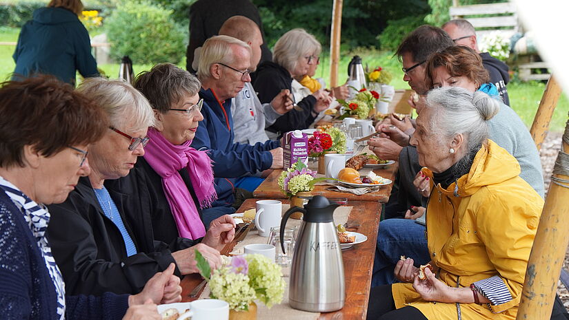 Ein lange Frühstückstafel unter einem Sonnensegel bei frischem Wetter: Menschen essen gemeinsam.
