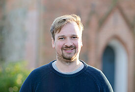 EIn junger Mann mit blonden Haaren, Bart und mit einem blauem Pullover gekleidet steht lächelnd vor einer Kirche.