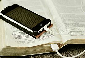 Smartphone mit Bibeltext liegt auf einer Bibel; das Kabel des Smartphones läuft in die Bücherseiten