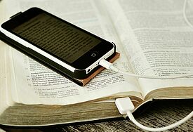 Smartphone mit Bibeltext liegt auf einer Bibel; das Kabel des Smartphones läuft in die Bücherseiten