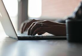 Hände tippen auf der Tastatur eines Laptops. Im Hintergrund bricht Licht durch ein Fenster in den Raum.