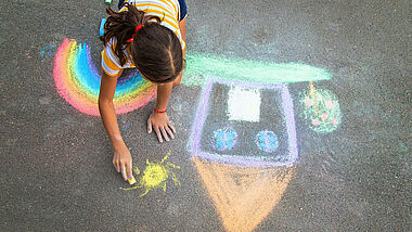 Ein Kind malt ein Haus und einen Regenbogen auf den Asphalt mit bunter Kreide.