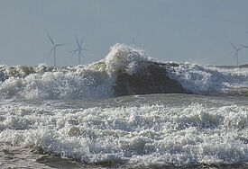 Nordseewellen rauschen an den Strand, im Hintergrund stehen Windräder im Meer.