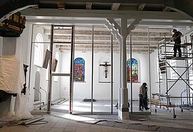 Ein Gerüst steht im inneren einer Kirche, Bauarbeiter stehen darauf und daneben.