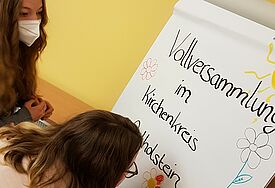 Zwei junge Frauen malen an einem Plakat. Neben bunten Blumen und einer gelben Sonne steht geschrieben: "Vollversammlung im Kirchenkreis Ostholstein".