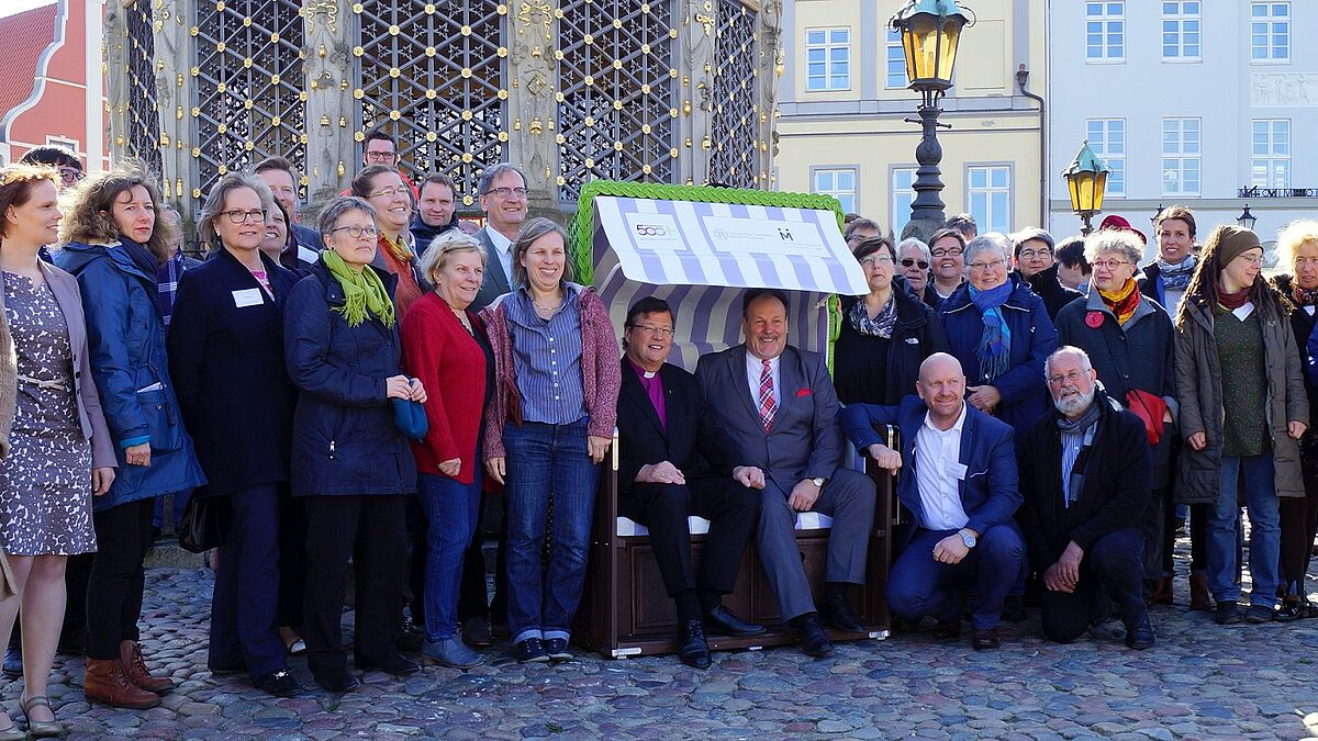 Bischof Dr. Hans-Jürgen Abromeit und Bernd Fischer, Geschäftsführer des Landestourismusverbandes Mecklenburg Vorpommern, werden während des Ausprobierens des Strandkorbs auf dem Marktplatz in Wismar von Teilnehmenden des Fachkongresses Kirche und Tourismus im Norden umringt