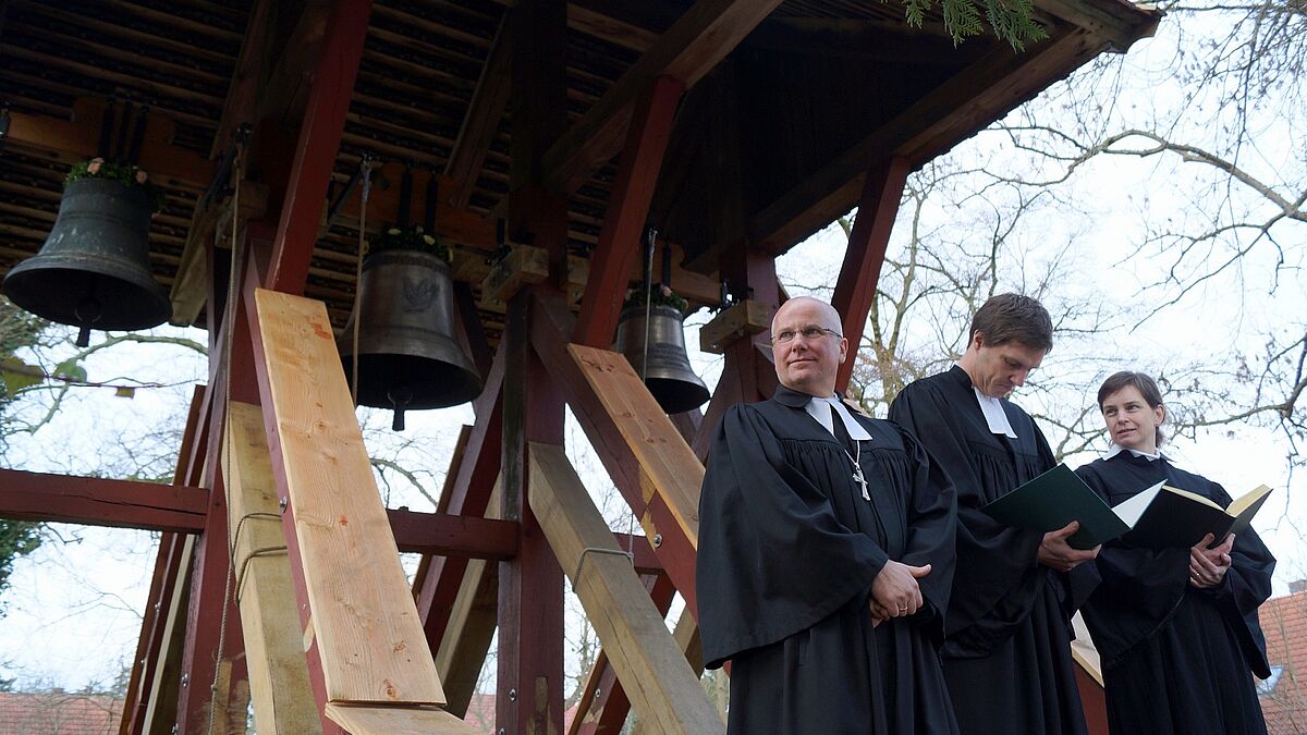 Propst Gerd Panknin (links), Pastor Christian Bauer und Pastorin Katrin Krüger vor den mit Blumen geschmückten Glocken - die historische Glocke hängt links, in der Mitte die neue "Friedensglocke" und rechts die Glocke für "Zuversicht und Stärke"