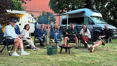 Im Kreis auf Stühlen auf dem Rasen sitzt die Moderatorin Bettina Tietjen (Mitte), die mit ihrem Wohnmobil Station bei Pastorin Silke Kühn in Kartlow (3.v.li) machte. Mit dabei waren die Promi-Camper und -Camperinnen Axel Schulz, Isabel Varell, Leo Reisinger, Ariana Baborie und Pascal Hens, genannt „Pommes“. Im Hintergrund die Kartlower Johanniskirche.