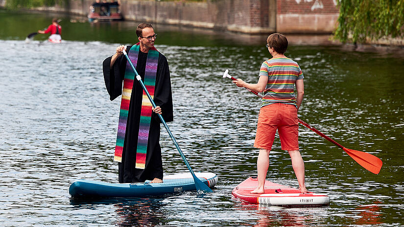 Pastor im Talar auf einen Stand-Up-Board auf der Hamburger Alster im Gespräch mit einem anderen Paddler.