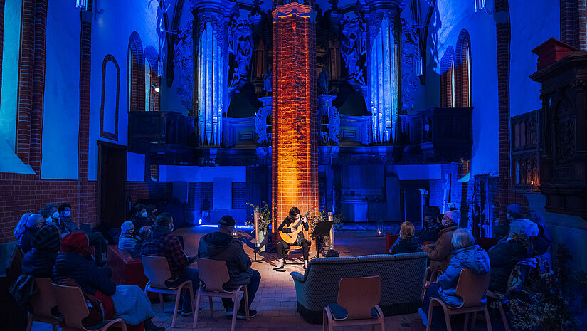 Eine dunkler Kirchenraum ist in blaues Licht getaucht. Besucherinnen und Besucher hören einer Gitarrenspielerin zu